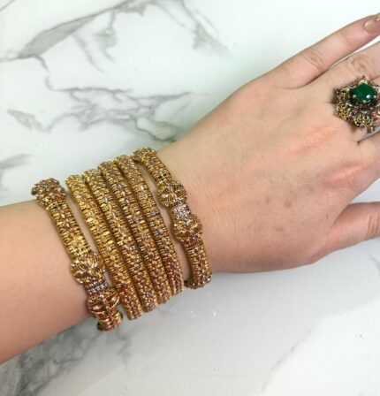 A hand wearing six gold-toned textured bangles with intricate detailing and floral designs, displayed on a white marble background