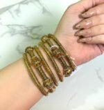 A hand wearing a set of antique gold-plated bangles with intricate beaded detailing and ornate dome-shaped accents, displayed against a marble background.