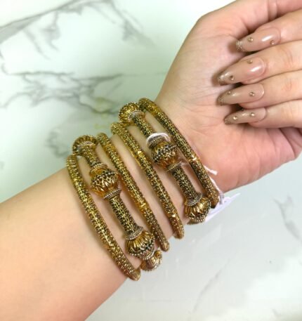 A hand wearing a set of antique gold-plated bangles with intricate beaded detailing and ornate dome-shaped accents, displayed against a marble background.