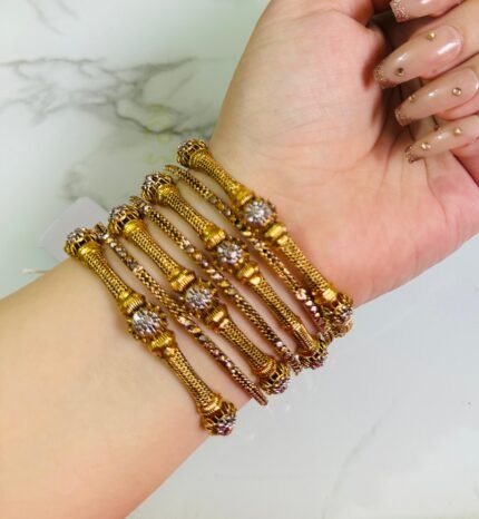 A hand wearing a set of six gold bangles with a fine mesh design and floral-shaped crystal embellishments. The bangles are lightweight, cylindrical, and elegant, set against a white marble background. A small tag is attached to one of the bangles with a string.