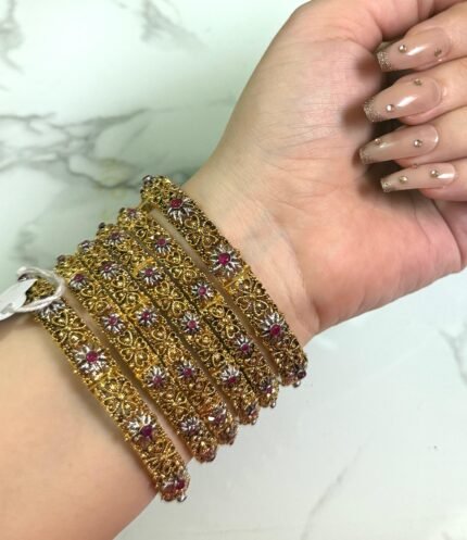 A hand adorned with six gold-plated bangles featuring intricate floral patterns and studded with red and silver stones, displayed against a light marble background.