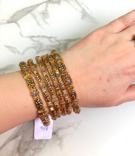A hand wearing a set of six intricately designed gold-plated bangles with detailed filigree and beadwork. The bangles feature a traditional Indian design