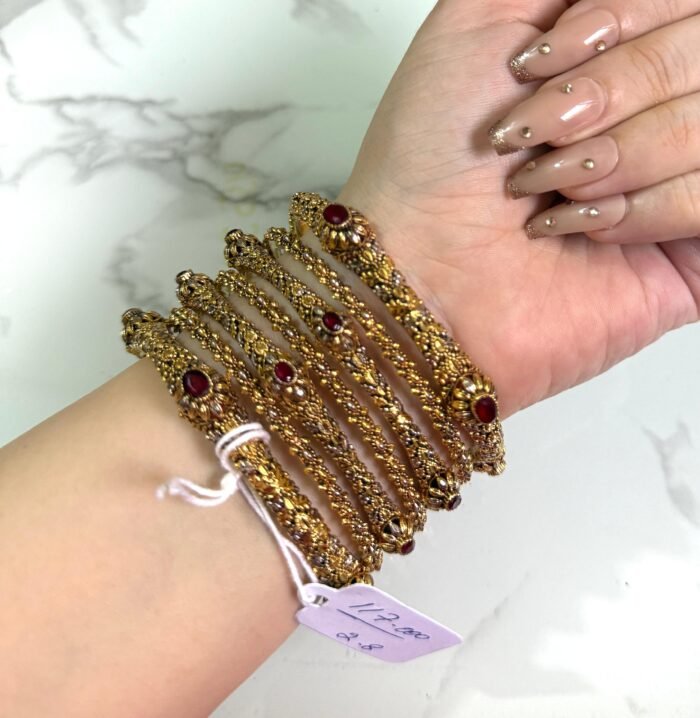 A hand adorned with a set of six antique gold bangles, each intricately designed with ornate patterns and embedded with oval-shaped deep red stones