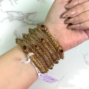 A hand adorned with a set of six antique gold bangles, each intricately designed with ornate patterns and embedded with oval-shaped deep red stones