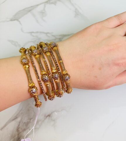 A hand wearing a set of six gold bangles with a fine mesh design and floral-shaped crystal embellishments. The bangles are lightweight, cylindrical, and elegant, set against a white marble background. A small tag is attached to one of the bangles with a string.