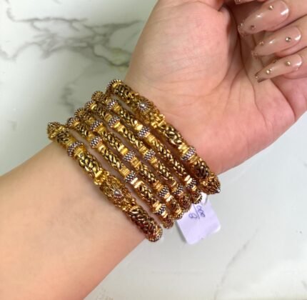 hand adorned with six intricately designed gold-plated bangles featuring braided patterns, antique-finish detailing, and small pearl-like bead embellishments. The bangles have a rich traditional look and are displayed against a white marble background. A price tag is attached to one of the bangles, and the nails are styled with nude polish and gold decorations