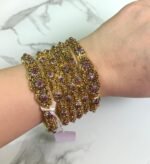 A hand wearing six antique gold bangles with intricate floral designs, featuring ruby red and white stone embellishments. The bangles are detailed with filigree work and displayed against a white marble background. A small price tag is tied to one bangle, and the overall style is traditional and ornate.