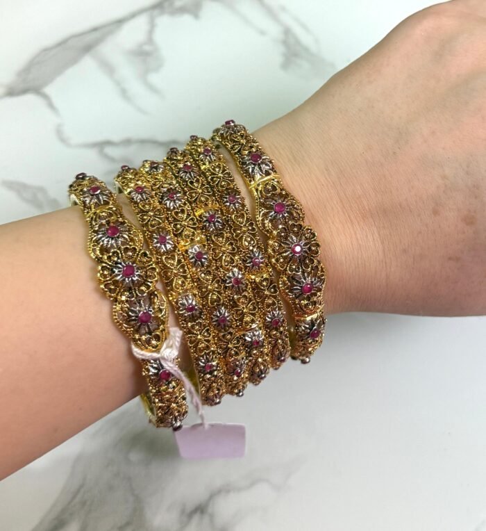 A hand wearing six antique gold bangles with intricate floral designs, featuring ruby red and white stone embellishments. The bangles are detailed with filigree work and displayed against a white marble background. A small price tag is tied to one bangle, and the overall style is traditional and ornate.
