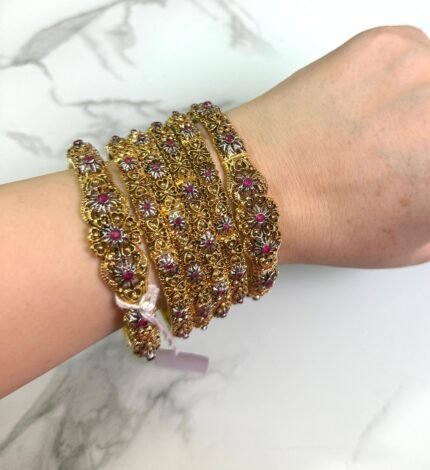 A hand wearing six antique gold bangles with intricate floral designs, featuring ruby red and white stone embellishments. The bangles are detailed with filigree work and displayed against a white marble background. A small price tag is tied to one bangle, and the overall style is traditional and ornate.