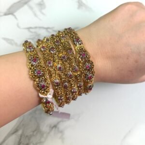 A hand wearing six antique gold bangles with intricate floral designs, featuring ruby red and white stone embellishments. The bangles are detailed with filigree work and displayed against a white marble background. A small price tag is tied to one bangle, and the overall style is traditional and ornate.