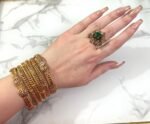 A wrist adorned with six intricately designed gold-plated mesh bangles featuring diamond-like stone clusters, set against a white marble background. The bangles showcase a braided texture and elegant beadwork, adding a luxurious and festive look.