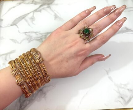 A wrist adorned with six intricately designed gold-plated mesh bangles featuring diamond-like stone clusters, set against a white marble background. The bangles showcase a braided texture and elegant beadwork, adding a luxurious and festive look.