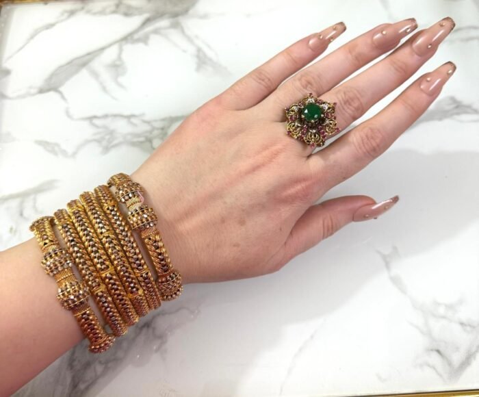 A wrist adorned with six intricately designed gold-plated mesh bangles featuring diamond-like stone clusters, set against a white marble background. The bangles showcase a braided texture and elegant beadwork, adding a luxurious and festive look.