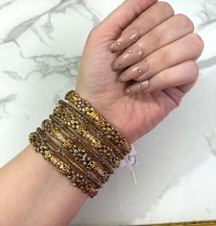 A wrist adorned with six intricately designed gold-plated bangles featuring floral motifs, beaded textures, and white stone embellishments, displayed against a white marble background.