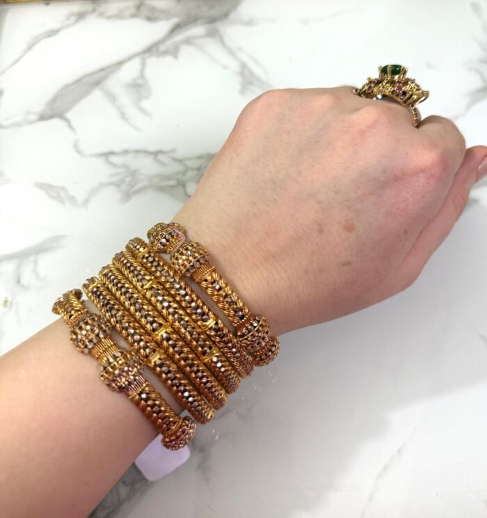 A wrist adorned with six intricately designed gold-plated mesh bangles featuring diamond-like stone clusters, set against a white marble background. The bangles showcase a braided texture and elegant beadwork, adding a luxurious and festive look.