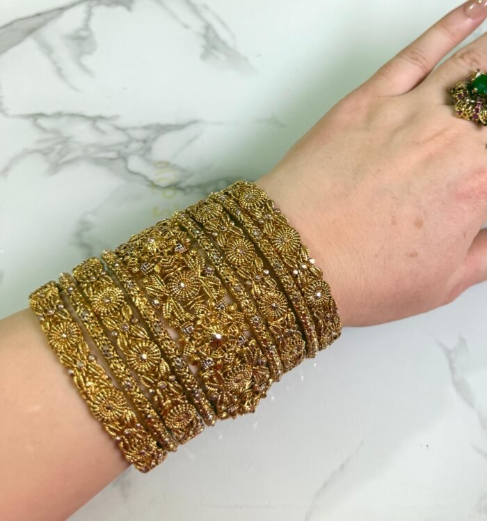A hand wearing eight antique gold bangles with detailed floral filigree work and subtle white embellishments, shown on a white marble background.