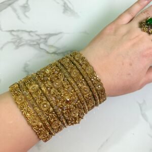 A hand wearing eight antique gold bangles with detailed floral filigree work and subtle white embellishments, shown on a white marble background.