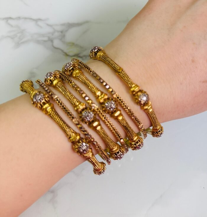 A hand wearing a set of six gold bangles with a fine mesh design and floral-shaped crystal embellishments. The bangles are lightweight, cylindrical, and elegant, set against a white marble background. A small tag is attached to one of the bangles with a string.