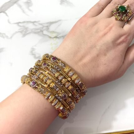 A wrist adorned with six ornate dual-tone gold bangles featuring ruby-colored stones, intricate detailing, and paired with a large green gemstone ring on a white marble background
