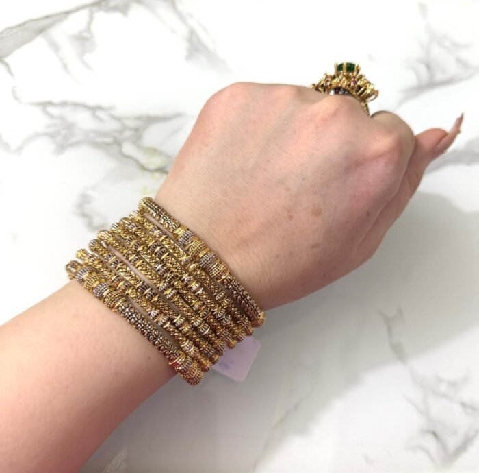 A hand wearing eight intricately detailed gold-tone bangles with dual-tone embellishments and a statement green gemstone ring, displayed on a white marble surface.