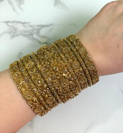 A hand wearing eight antique gold bangles with detailed floral filigree work and subtle white embellishments, shown on a white marble background.