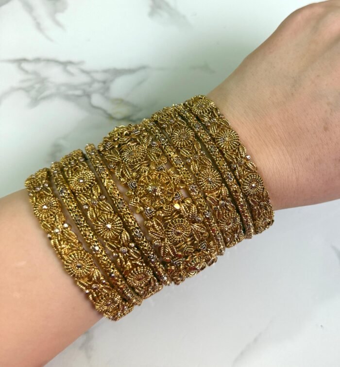 A hand wearing eight antique gold bangles with detailed floral filigree work and subtle white embellishments, shown on a white marble background.