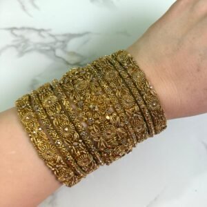 A hand wearing eight antique gold bangles with detailed floral filigree work and subtle white embellishments, shown on a white marble background.