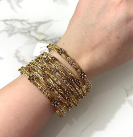 A hand adorned with eight gold bangles showcasing intricate chevron engravings and shimmering stone embellishments, set against a marble background.