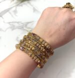 A wrist adorned with six ornate dual-tone gold bangles featuring ruby-colored stones, intricate detailing, and paired with a large green gemstone ring on a white marble background.