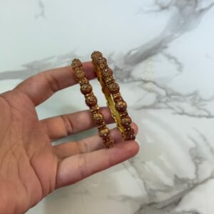 A pair of intricately designed antique copper-toned bangles with dome-shaped bead detailing, held in hand against a white marble background with a gold-edged tray.
