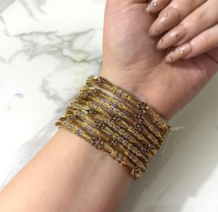 A hand adorned with eight gold bangles showcasing intricate chevron engravings and shimmering stone embellishments, set against a marble background.