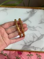 A close-up of a hand holding two antique-style gold bangles, featuring delicate maroon stone embellishments and detailed filigree patterns, displayed against a white marble background and pink floral fabric. Ideal for traditional or festive occasions.