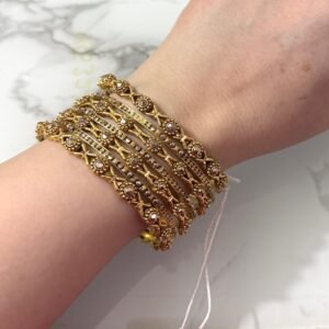 A hand adorned with eight intricately designed antique gold bangles featuring cross motifs and embedded pearl-like beads, displayed on a marble surface.