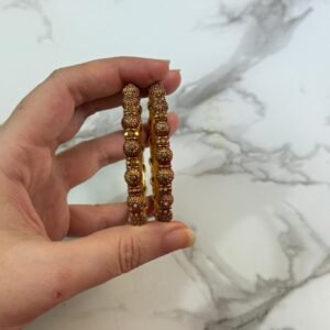 A pair of intricately designed antique copper-toned bangles with dome-shaped bead detailing, held in hand against a white marble background with a gold-edged tray.