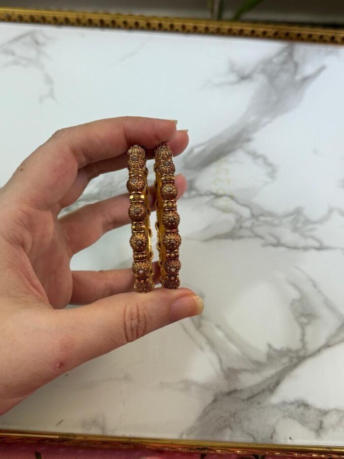A pair of intricately designed antique copper-toned bangles with dome-shaped bead detailing, held in hand against a white marble background with a gold-edged tray.