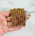 A hand holding six antique gold-plated bangles adorned with detailed floral and leaf motifs, and studded with sparkling white stones. The intricate design and craftsmanship are highlighted against a white marble background.