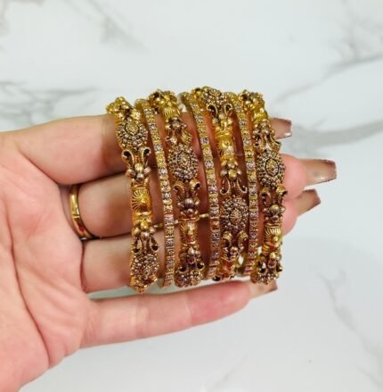 A hand holding six antique gold-plated bangles adorned with detailed floral and leaf motifs, and studded with sparkling white stones. The intricate design and craftsmanship are highlighted against a white marble background.