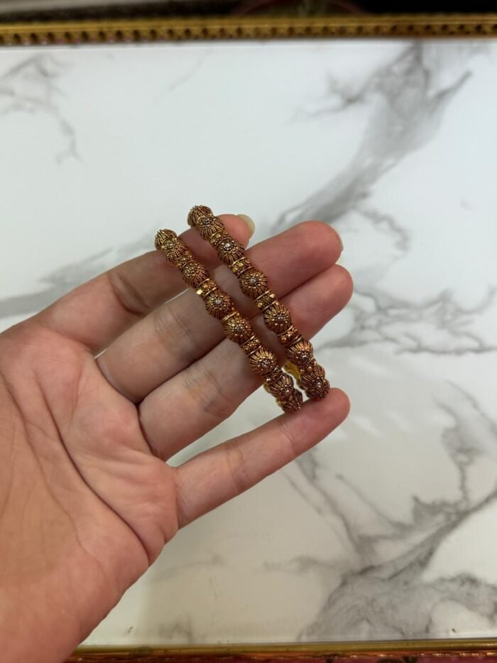 A pair of intricately designed antique copper-toned bangles with dome-shaped bead detailing, held in hand against a white marble background with a gold-edged tray.