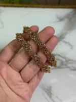 Close-up of intricately designed antique gold bangles held in hand, featuring floral filigree patterns and small pink gemstone embellishments.