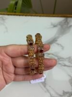 Hand holding a pair of traditional gold bangles with detailed textured design and oval red gemstones, placed against a marble background.