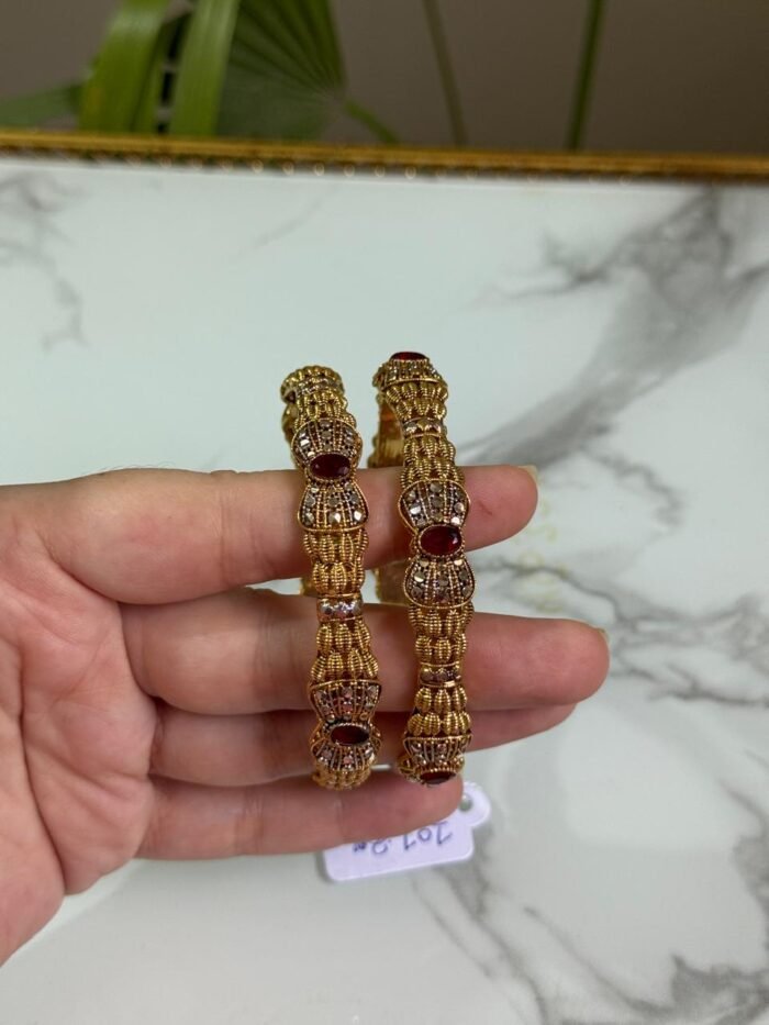 Hand holding a pair of traditional gold bangles with detailed textured design and oval red gemstones, placed against a marble background.