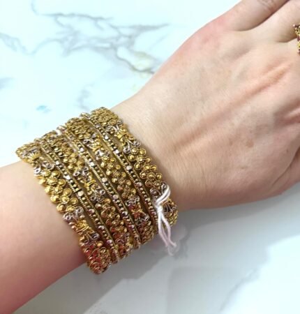 A wrist adorned with eight gold-plated bangles featuring floral cluster patterns and silver-toned accents, displayed on a white marble background