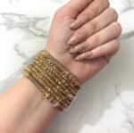 A hand wearing eight intricately detailed gold-tone bangles with dual-tone embellishments and a statement green gemstone ring, displayed on a white marble surface.
