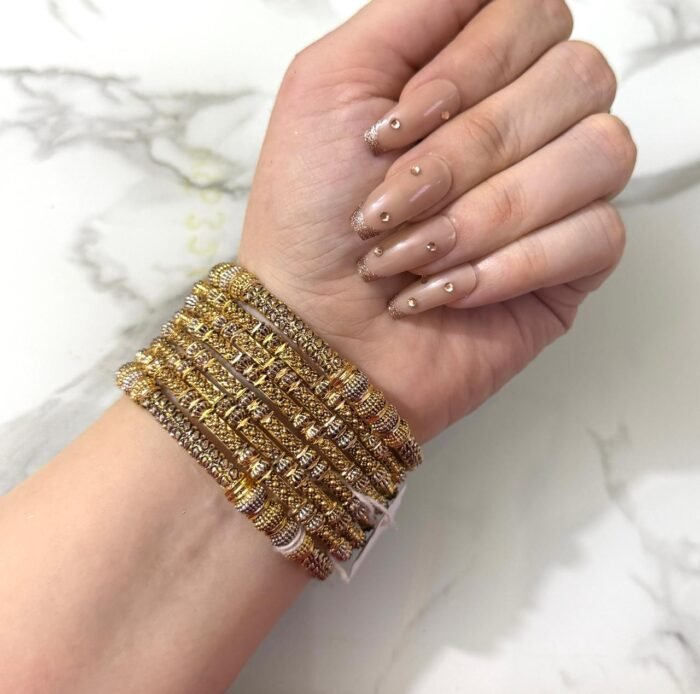 A hand wearing eight intricately detailed gold-tone bangles with dual-tone embellishments and a statement green gemstone ring, displayed on a white marble surface.