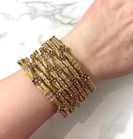A hand adorned with eight gold bangles showcasing intricate chevron engravings and shimmering stone embellishments, set against a marble background.