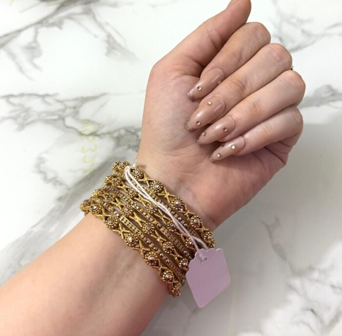 A hand adorned with eight intricately designed antique gold bangles featuring cross motifs and embedded pearl-like beads, displayed on a marble surface.