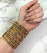 A hand wearing eight antique gold-plated bangles adorned with clustered white stones and floral patterns, displayed on a marble surface with manicured nails