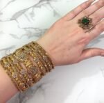 A hand wearing eight antique gold-plated bangles adorned with clustered white stones and floral patterns, displayed on a marble surface with manicured nails