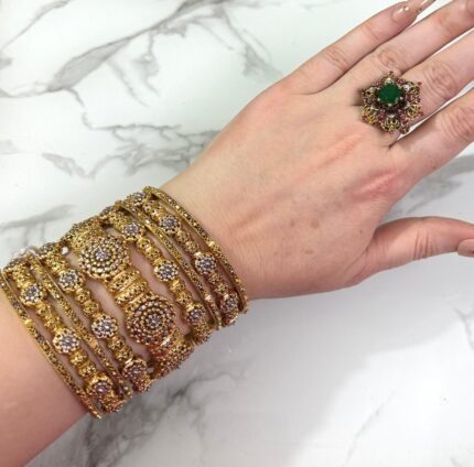 A hand wearing eight antique gold-plated bangles adorned with clustered white stones and floral patterns, displayed on a marble surface with manicured nails