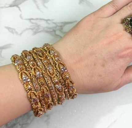 A hand wearing six intricately designed gold bangles featuring floral patterns adorned with small white stones. The bangles have a detailed leaf and petal motif throughout and are displayed against a white marble background. A tag with a price is tied to one bangle, and the nails are styled in nude polish with gold bead accents.